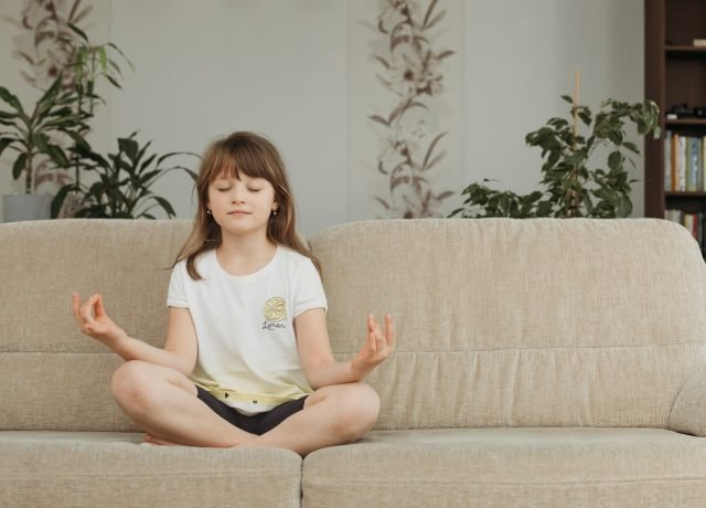 A cute girl schoolgirl is sitting at home on the couch and meditating. Children's yoga, meditation,